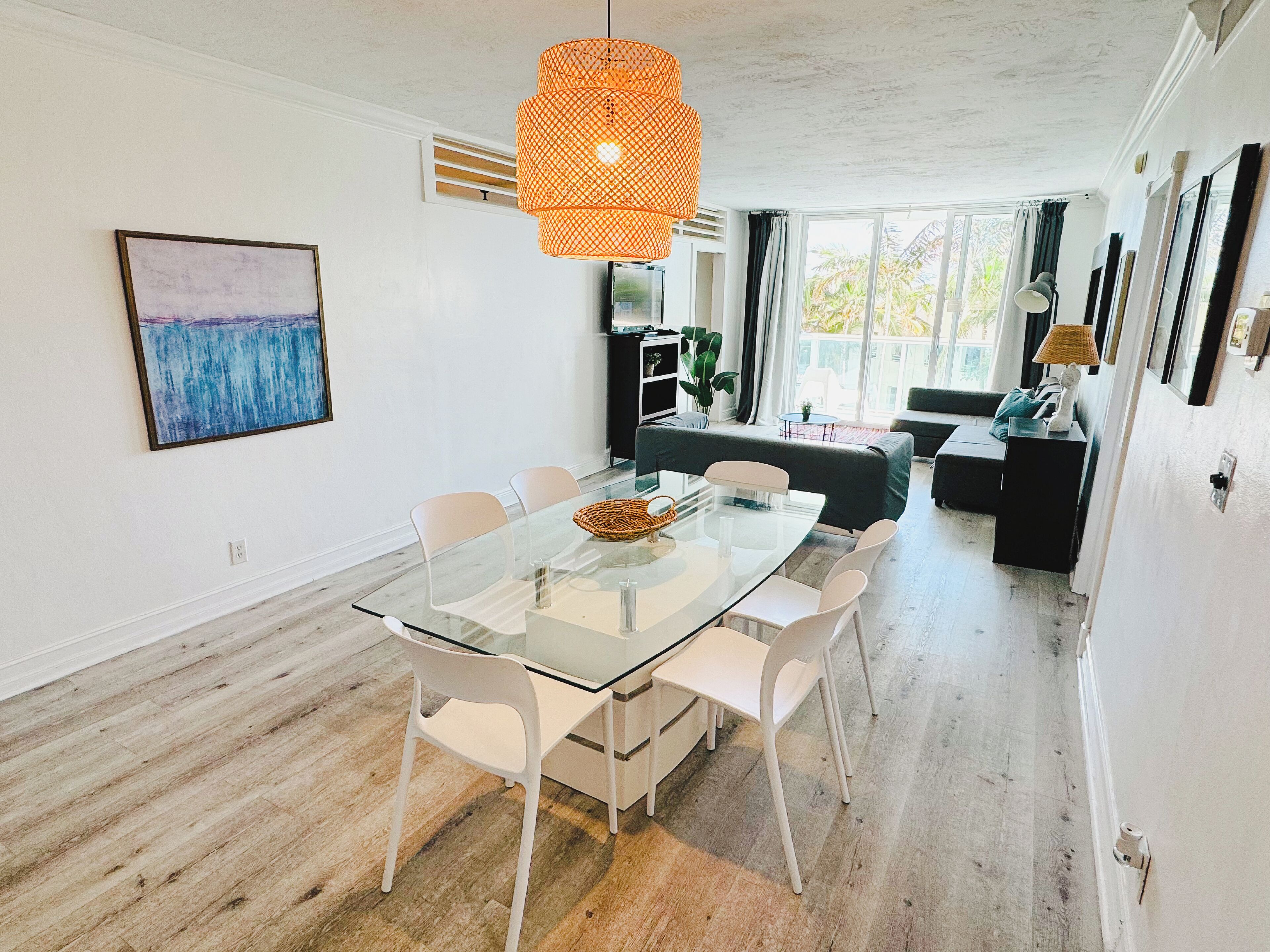 Una sala de estar moderna con una mesa de vidrio, sillas blancas, un sofá y un ventanal con vistas a palmeras.