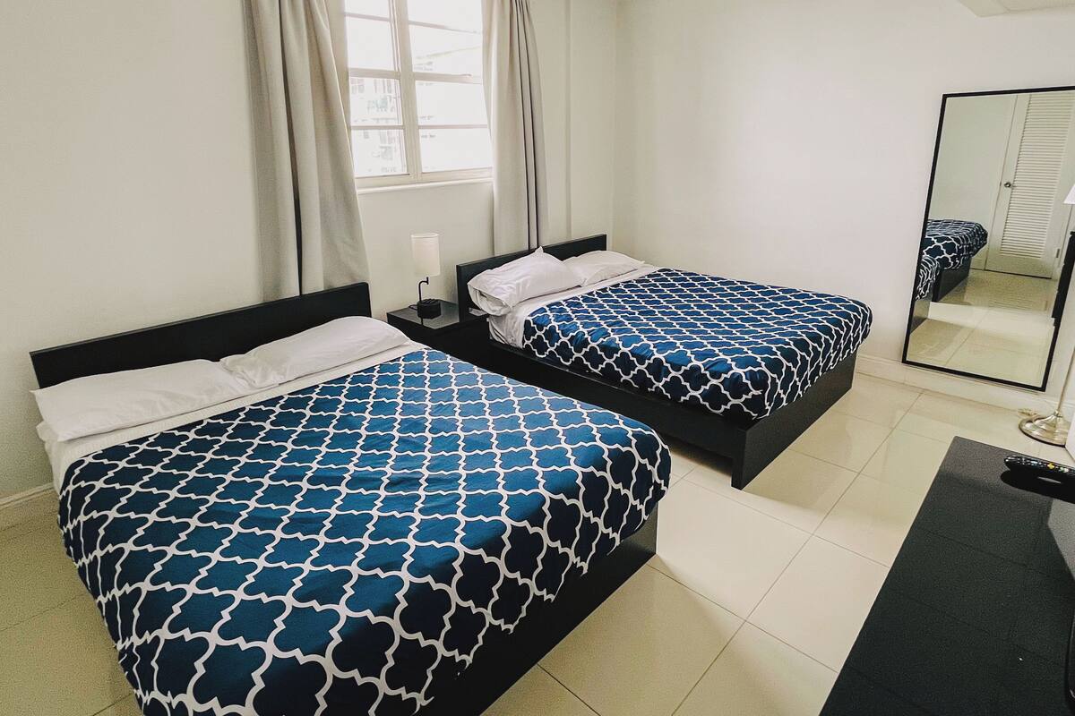 A hotel room with two beds, a window with curtains, a bedside table with a lamp, and a mirror on the wall.