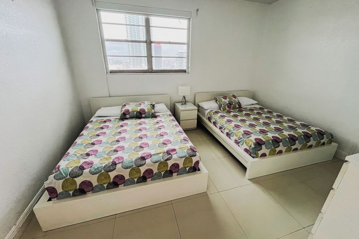 A small, clean room with two beds, a window, and a nightstand.