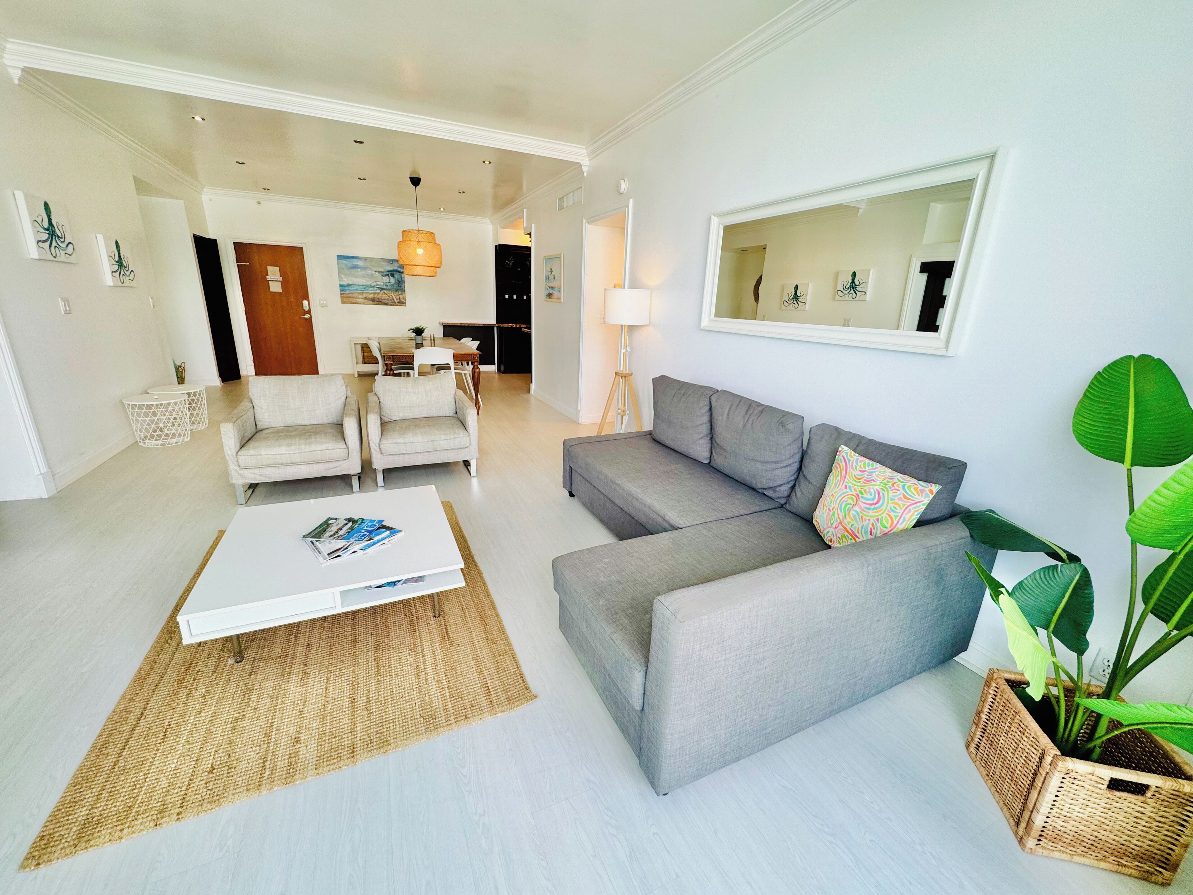A modern living room with a grey sofa, a white coffee table, and a large mirror. There is a dining area in the background with a table and chairs.