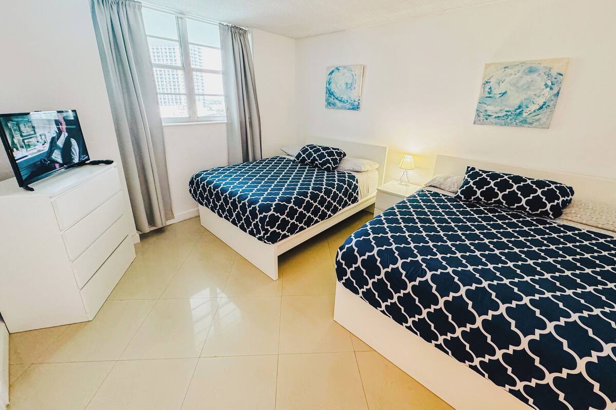 A hotel room with two beds, a TV, a desk, and a window with curtains.