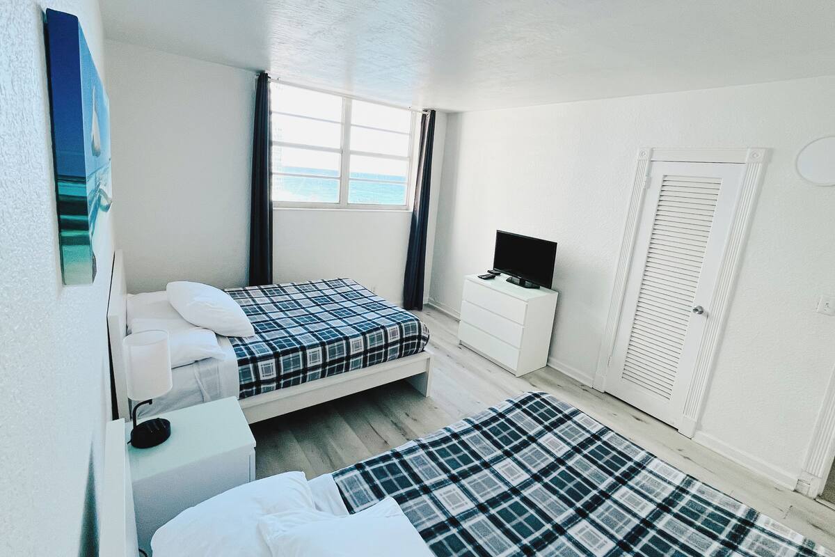 A bedroom with a bed, a TV on a dresser, a window with curtains, and a door.