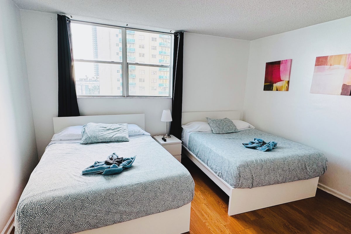 A twin room with two beds, a window with curtains, a bedside table with a lamp, and a wall with two paintings.
