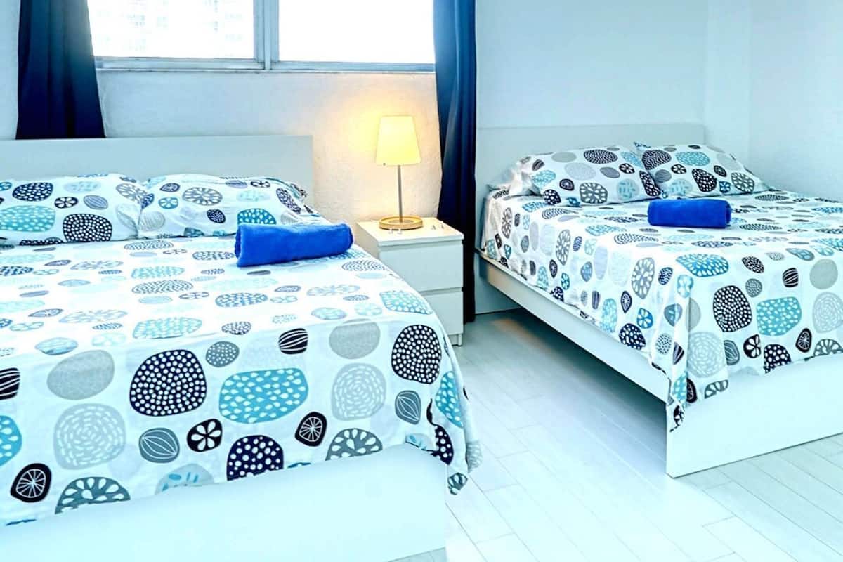 A hotel room with two beds, each with a patterned bedspread and a blue pillow. There is a bedside table with a lamp and a window with curtains.