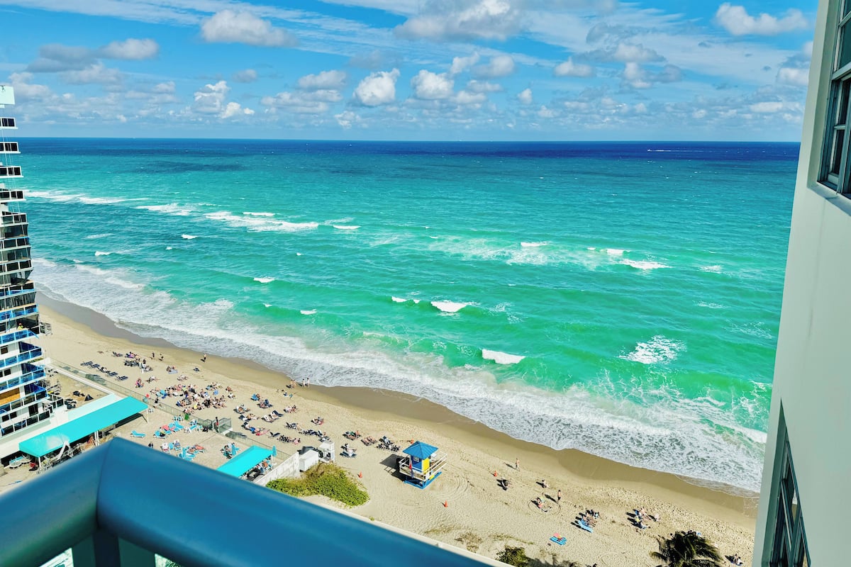 A beach with clear turquoise waters, a sandy shore, and a building with a balcony overlooking the scene.