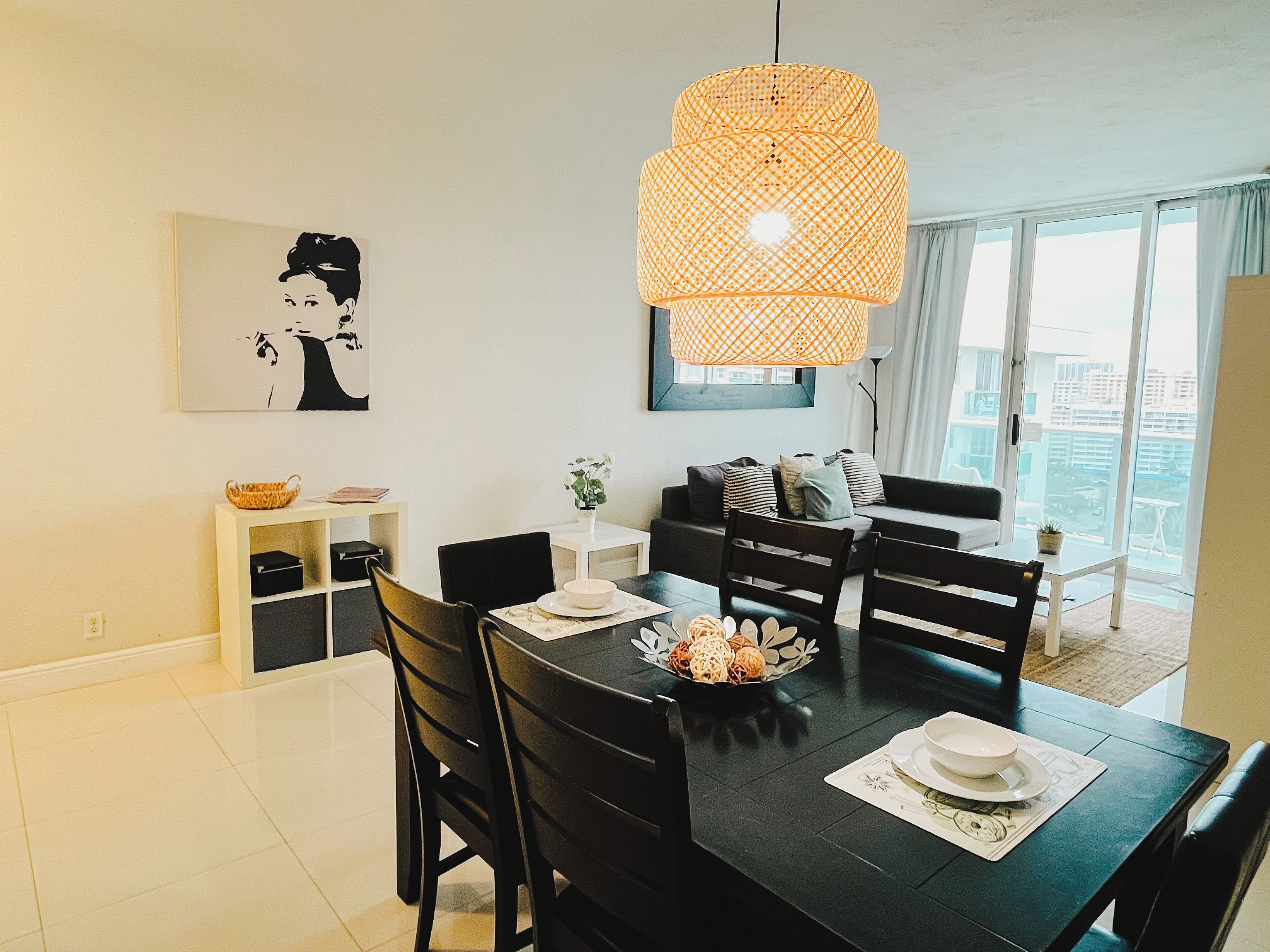 A dining area with a black table, white chairs, and a woven pendant light. There is a white shelf with a basket and a framed picture on the wall.