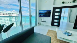 A modern living room with a large window overlooking a beach and ocean, a flat-screen TV mounted on the wall, a small table with magazines, and a comfortable sofa. - The Tides on Hollywood (Hollywood)
