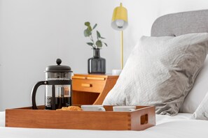 Uma bandeja de madeira com uma prensa francesa, um croissant e uma xícara, colocada sobre uma cama com um travesseiro e uma luminária ao fundo.