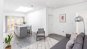 A modern living room with a grey sofa, a dining area with a kitchen island, and a large window providing natural light. - Beautiful Apartment in Central Neighbourhood (Cardiff)