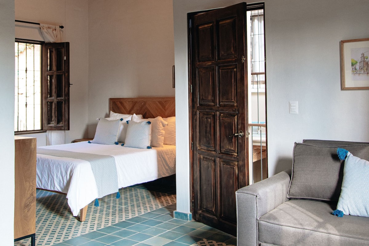 A bedroom with a bed, a wooden door, a grey sofa, and patterned floor tiles.