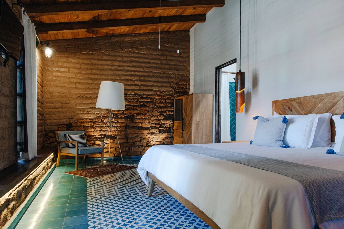 A bedroom with a stone wall, a bed with white bedding, a hanging lamp, and a chair with a striped cushion.