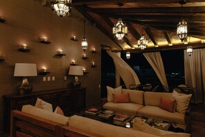 Un coin salon confortable avec un canapé, une table basse avec des livres, des lampes fixées au mur et une vue par une fenêtre agrémentée de rideaux.