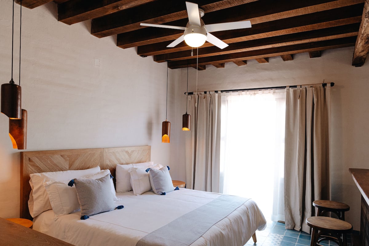 A bedroom with a wooden ceiling, a bed with white bedding, two hanging pendant lights, and a window with curtains.