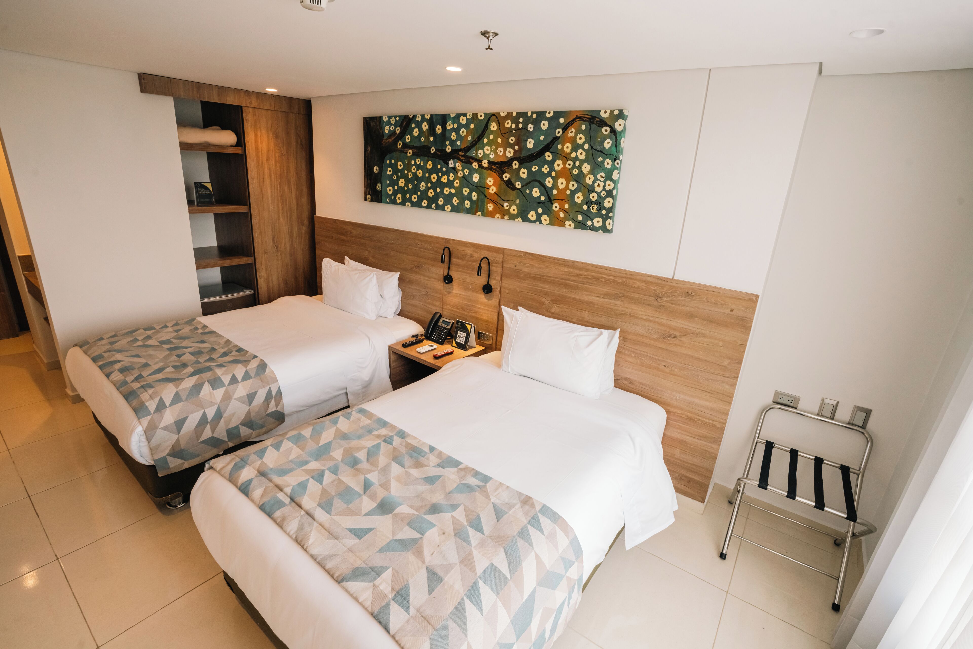 Een hotelkamer met twee bedden, een houten hoofdbord, een nachtkastje, een telefoon en een metalen rek.