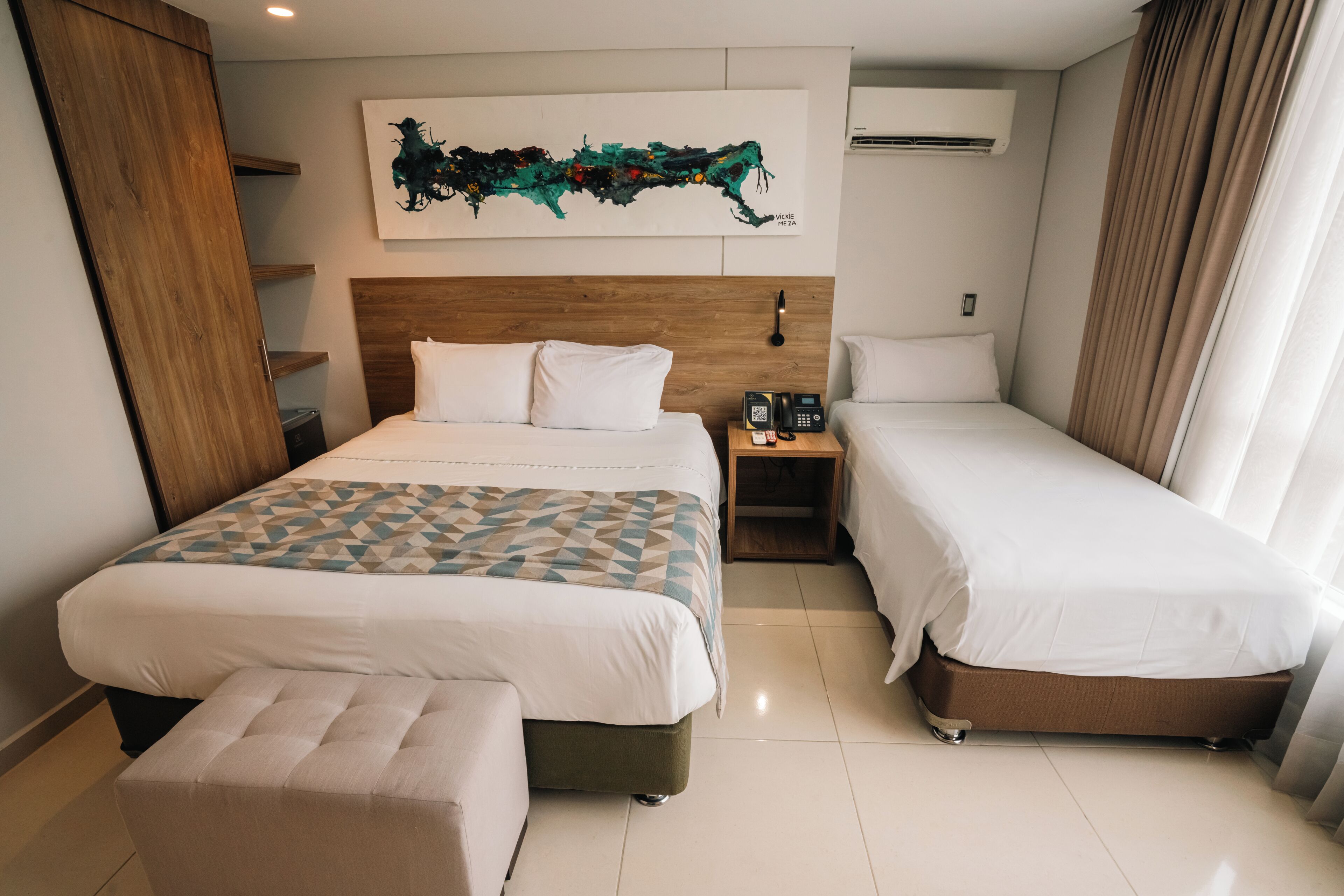 Une chambre d’hôtel avec un grand lit, un lit plus petit, une table de chevet avec un téléphone, une œuvre d’art accrochée au mur et une armoire en bois.