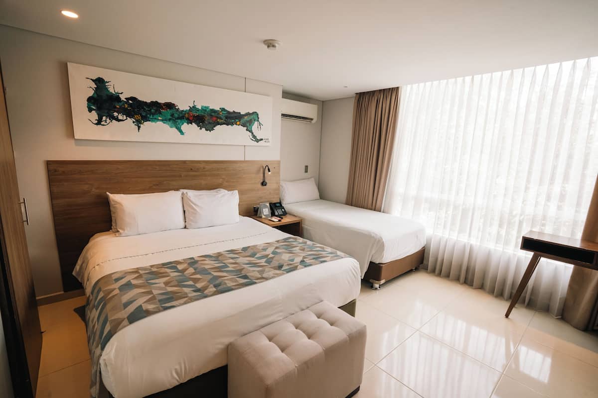 A hotel room with a large bed, a smaller bed, a bench, a desk, a phone, and a wall-mounted artwork.