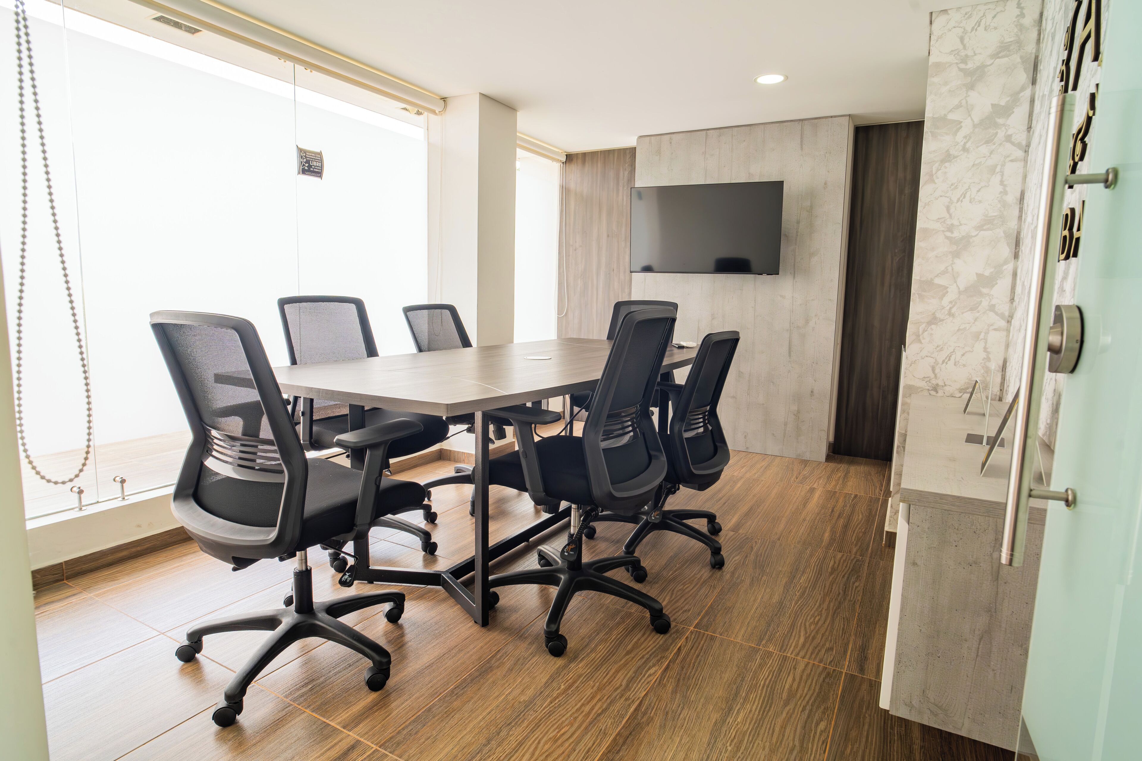 A conference room with a long table and six chairs, a flat-screen TV on the wall, and a wooden floor.