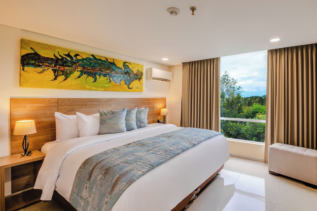 A hotel room with a large bed, a nightstand, a lamp, a painting on the wall, and a window with curtains.