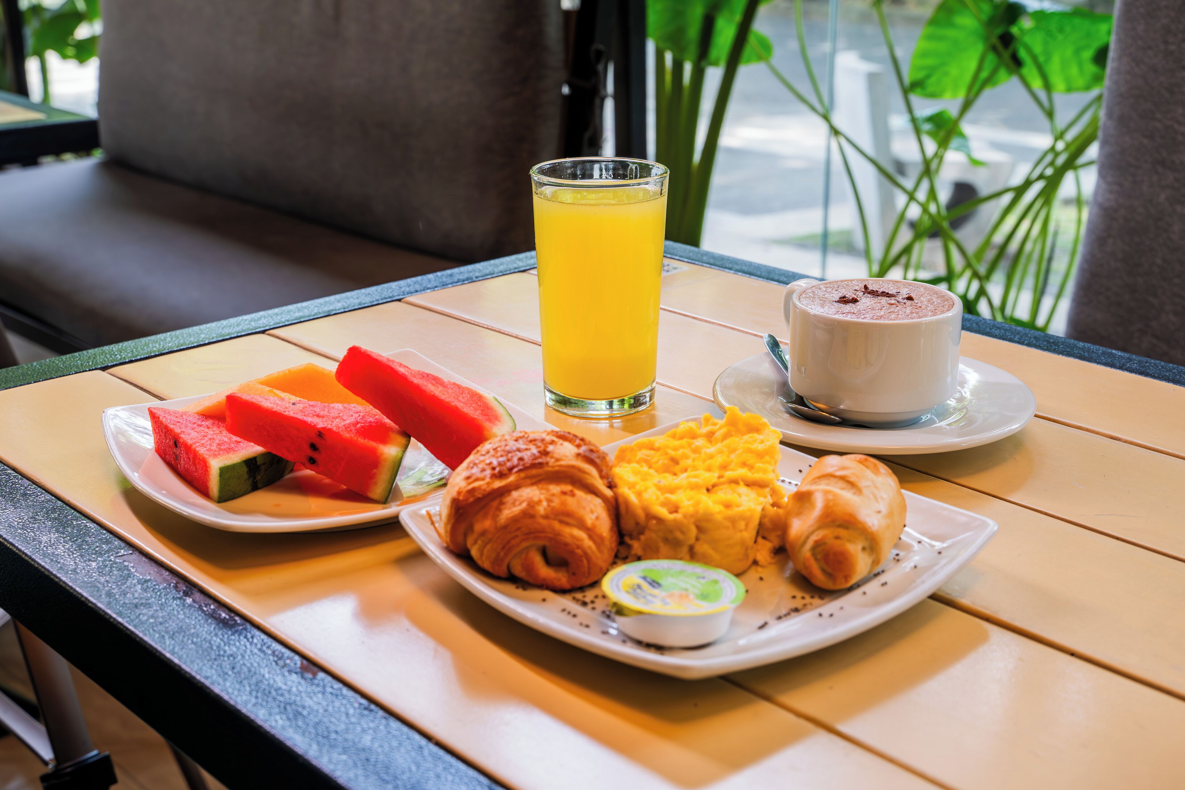 Un plateau de petit-déjeuner avec de la pastèque, des croissants, des œufs brouillés, un verre de jus d’orange et une tasse de café, posé sur une table en bois.