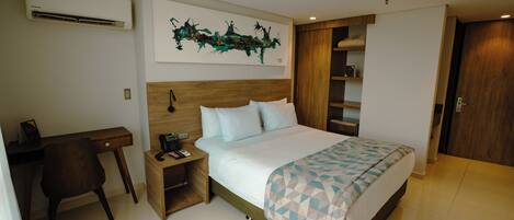 A hotel room with a bed, a desk, a chair, a nightstand, a wall-mounted air conditioner, and a wooden wardrobe.