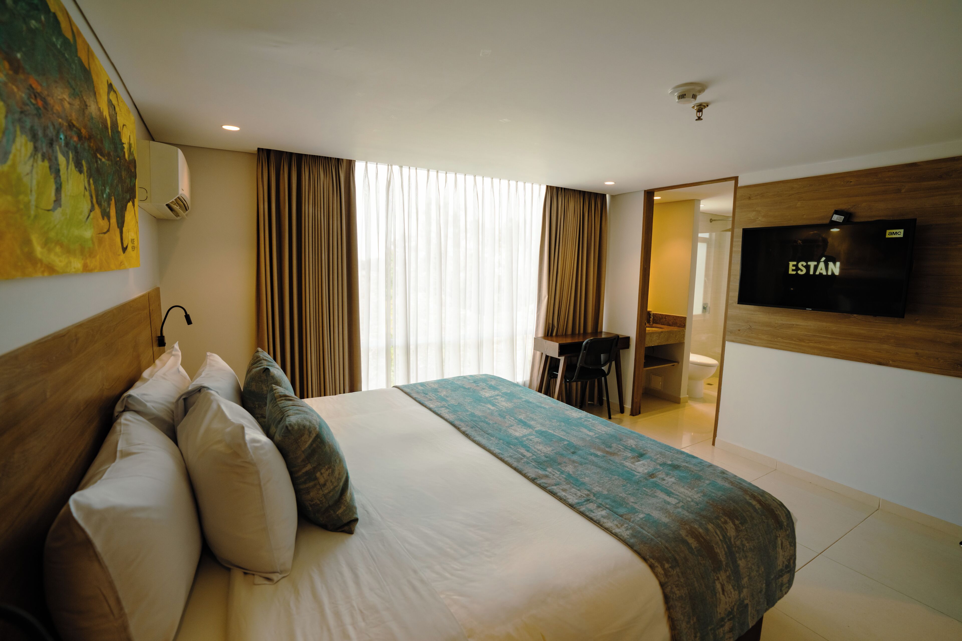 A hotel room with a bed, a desk, a television, and a large window with curtains.