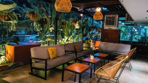 Un espacio lounge moderno con sofá, sillones y mesas, rodeado de plantas de interior y un amplio ventanal con vistas a una calle concurrida.