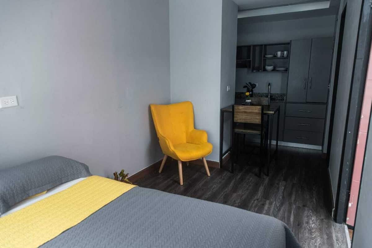 A compact hotel room with a bed, a yellow chair, and a kitchenette in the background.
