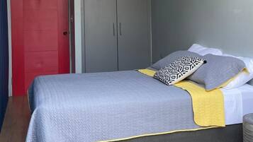 Un dormitorio con cama, ropa de cama gris y amarilla, una puerta roja, armario gris y un proyector montado en el techo.