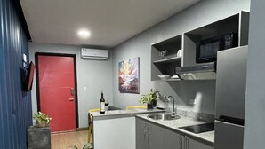 A modern kitchen with grey cabinets, a stainless steel sink, and a built-in microwave. There is a red door in the background and a painting on the wall. - Nomada Container Hotel Panama City (Panama City)