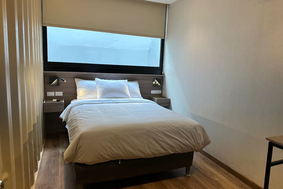 A modern bedroom with a large window, a wooden headboard, a bedside table, and a bed with white bedding.