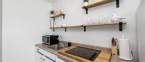 Una cucina moderna con mobili bianchi, forno a microonde integrato, fornello e mensole in legno sopra con bicchieri e una pianta.