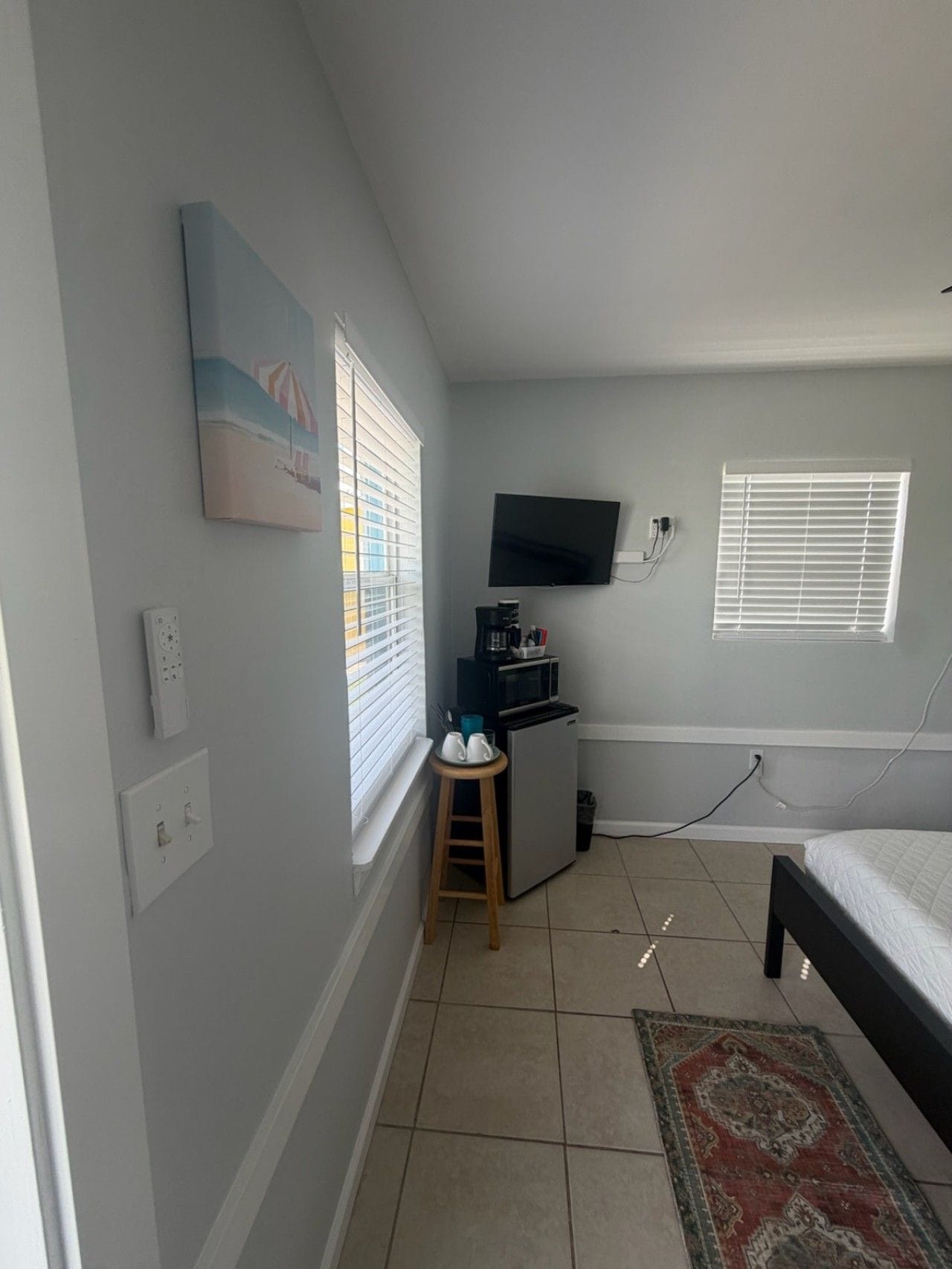 Una habitación pequeña y bien iluminada, con una cama, un televisor en la pared, una kitchenette y una ventana con persianas.