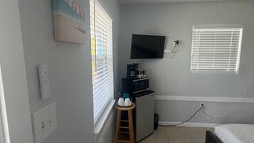 Una habitación pequeña y bien iluminada, con una cama, un televisor en la pared, una kitchenette y una ventana con persianas.