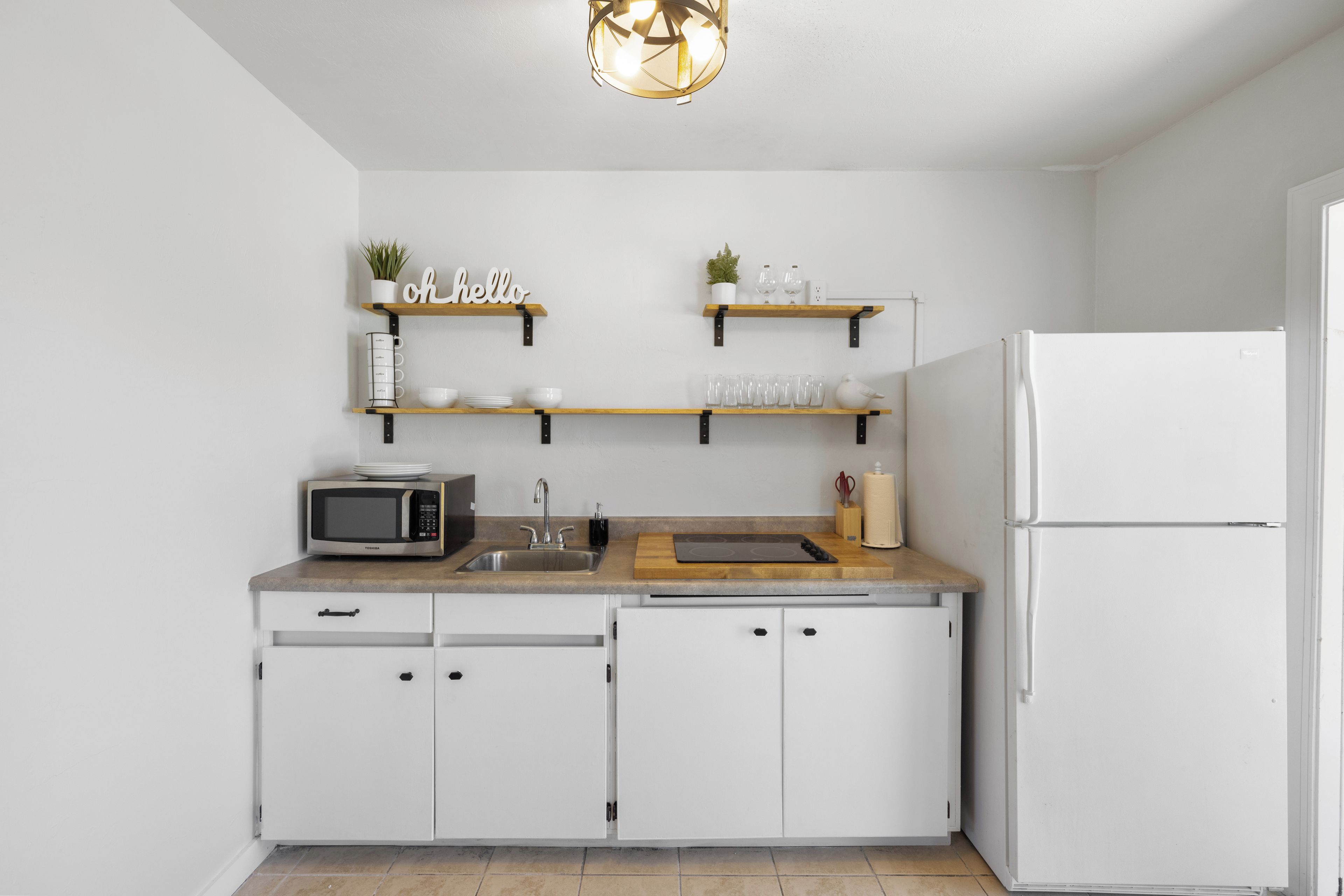 A modern kitchen with white cabinetry, a built-in microwave, a sink, and a refrigerator. There are wooden shelves above the countertop holding decorative items.