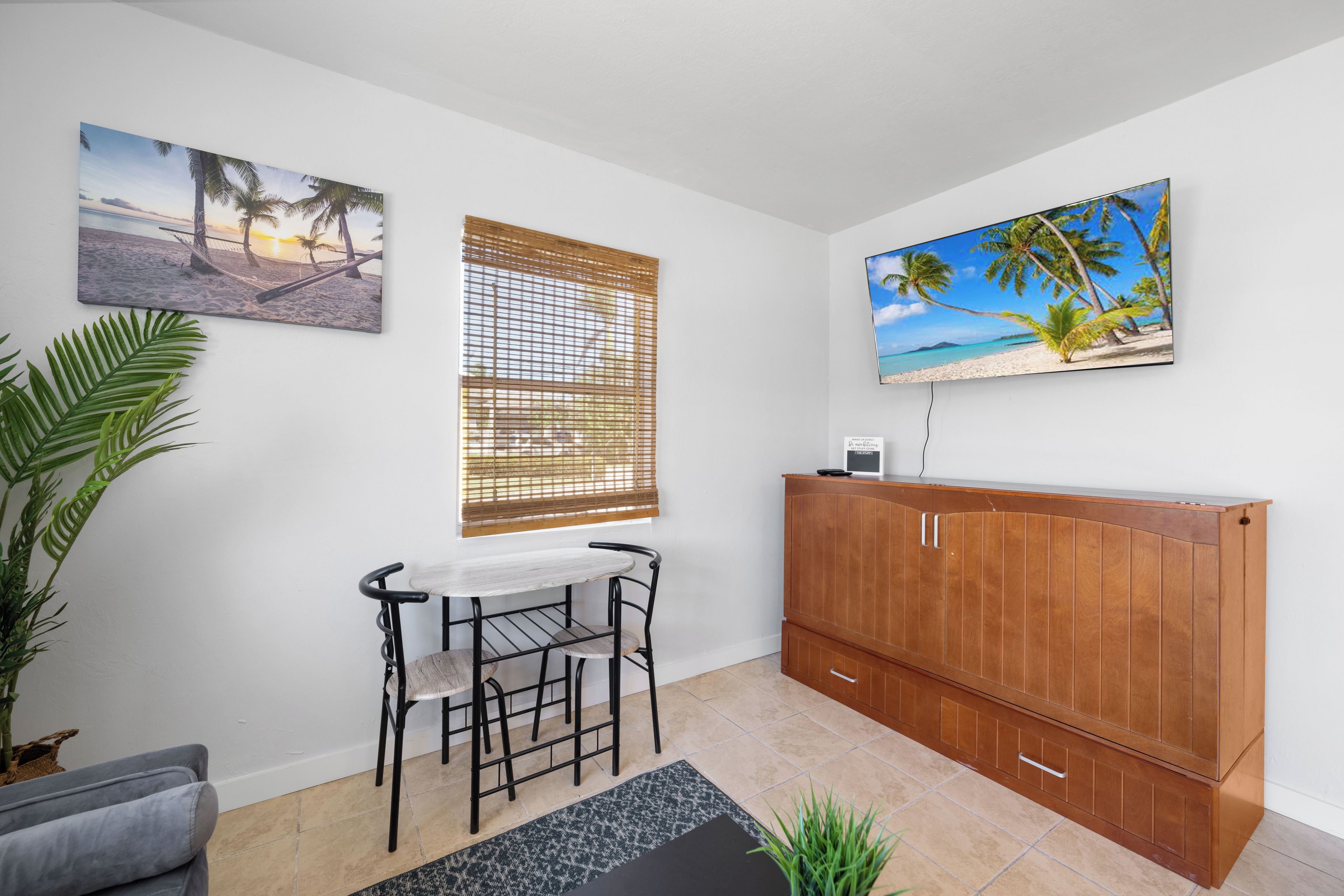 Una habitación con un armario de madera, una mesa con sillas, un ventanal con persianas y dos cuadros en la pared.