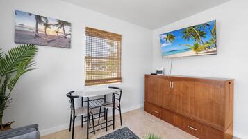 Una habitación con un armario de madera, una mesa con sillas, un ventanal con persianas y dos cuadros en la pared.