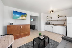 A modern living room with a wooden cabinet, a flat-screen TV, and a kitchen with white cabinets and a built-in microwave.