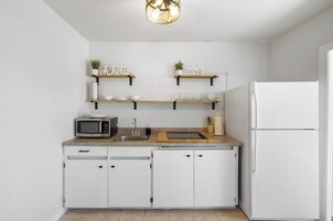 Una cocina moderna con armarios blancos, microondas integrado, pileta y refrigerador. Hay repisas de madera sobre la encimera con objetos decorativos.