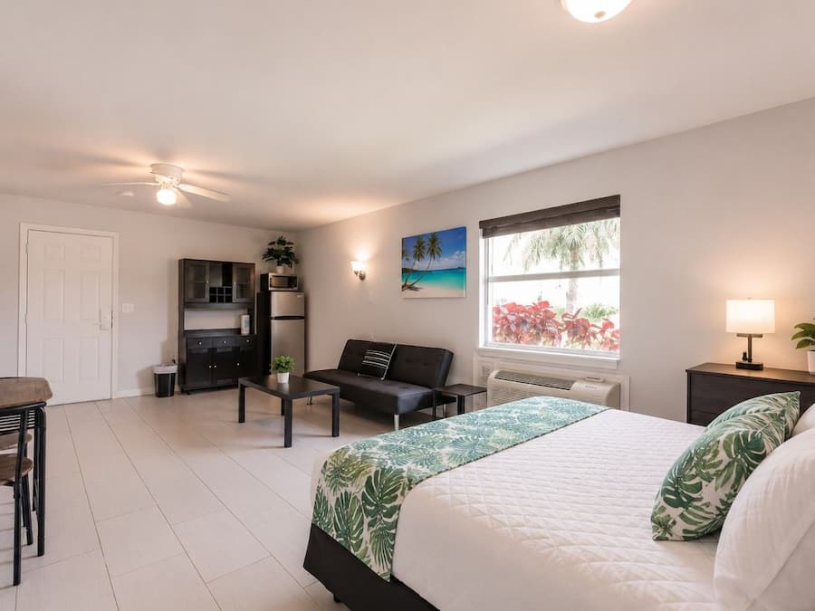 A modern bedroom with a bed, a desk, a sofa, a kitchenette, and a window with a view.