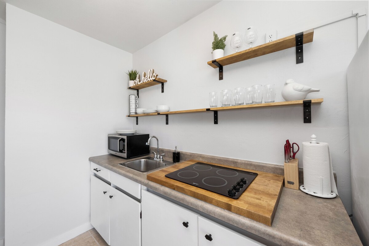 A modern kitchen with white cabinetry, a built-in microwave, a stove, and wooden shelves above holding glassware and a plant.