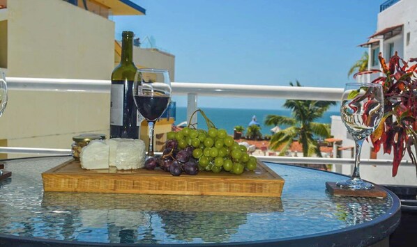 A table with a bottle of wine, a glass of red wine, a plate of cheese, and grapes, set on a balcony overlooking the sea.
