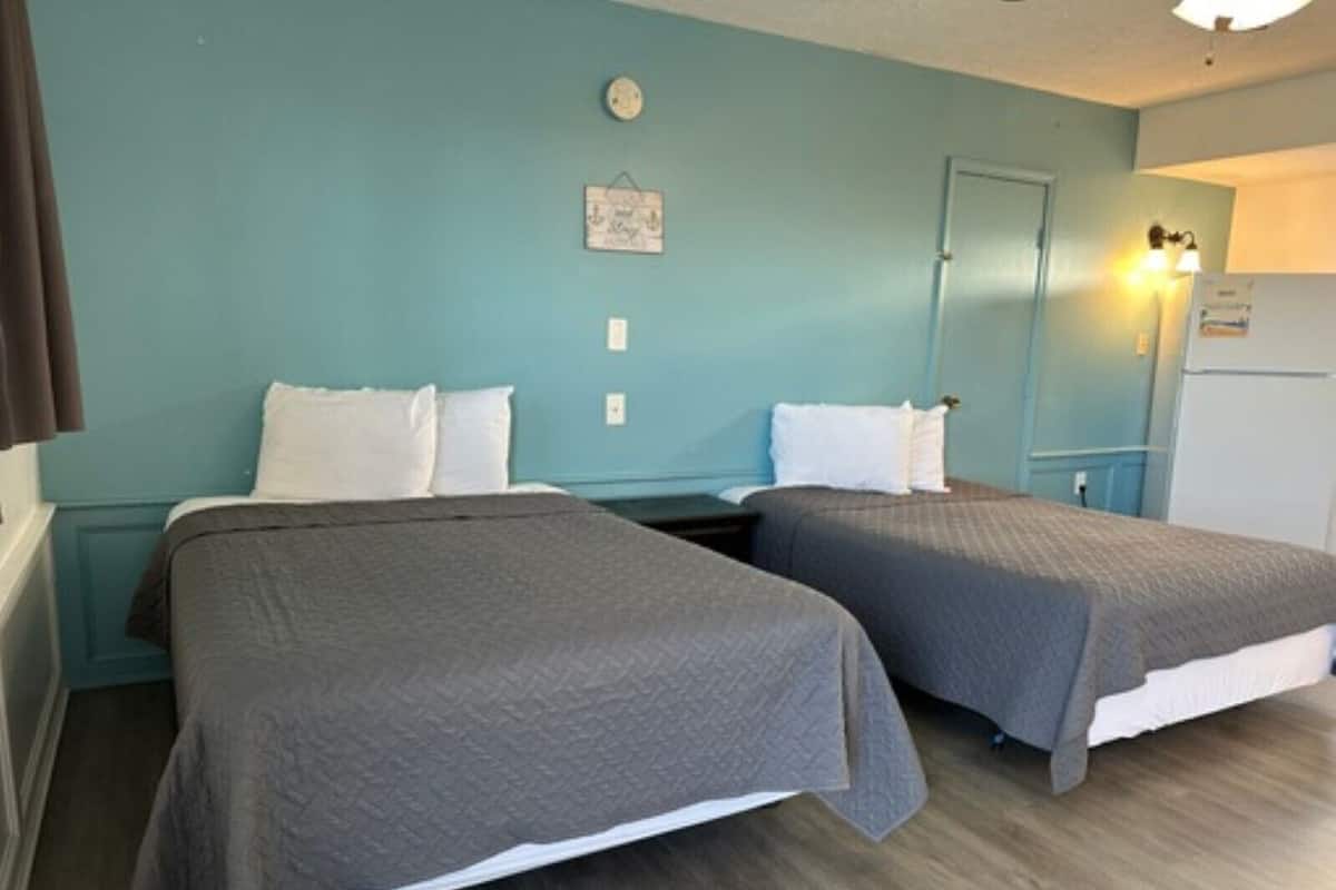 A room with two beds, teal walls, a small clock, and a door with a light above it.