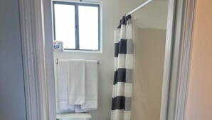 A bathroom with a toilet, a shower with a black and white striped curtain, and a window above the toilet. - Nautical Motel (Hampton)