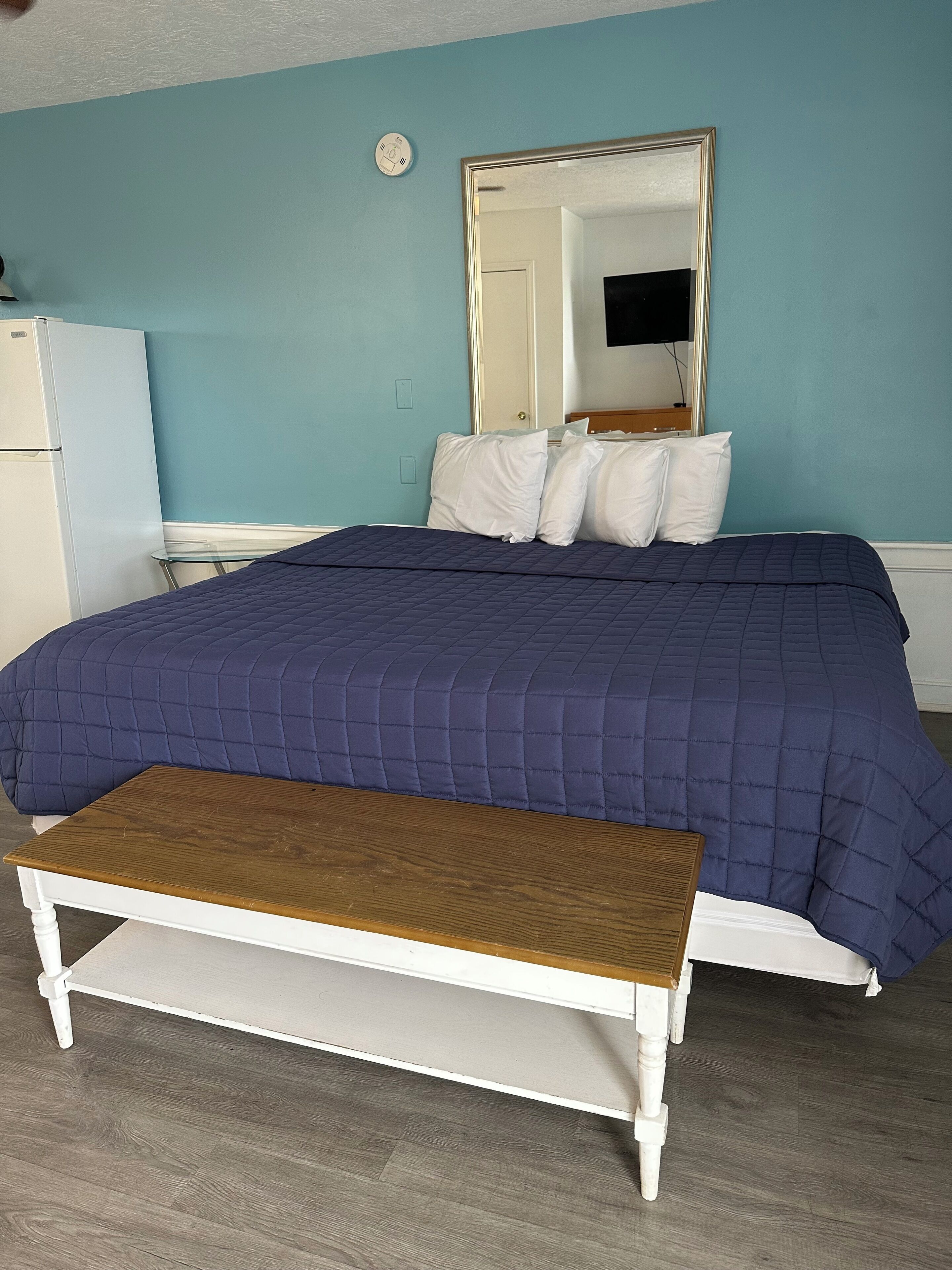 A bedroom with a bed, a bench, a mirror, and a white refrigerator.