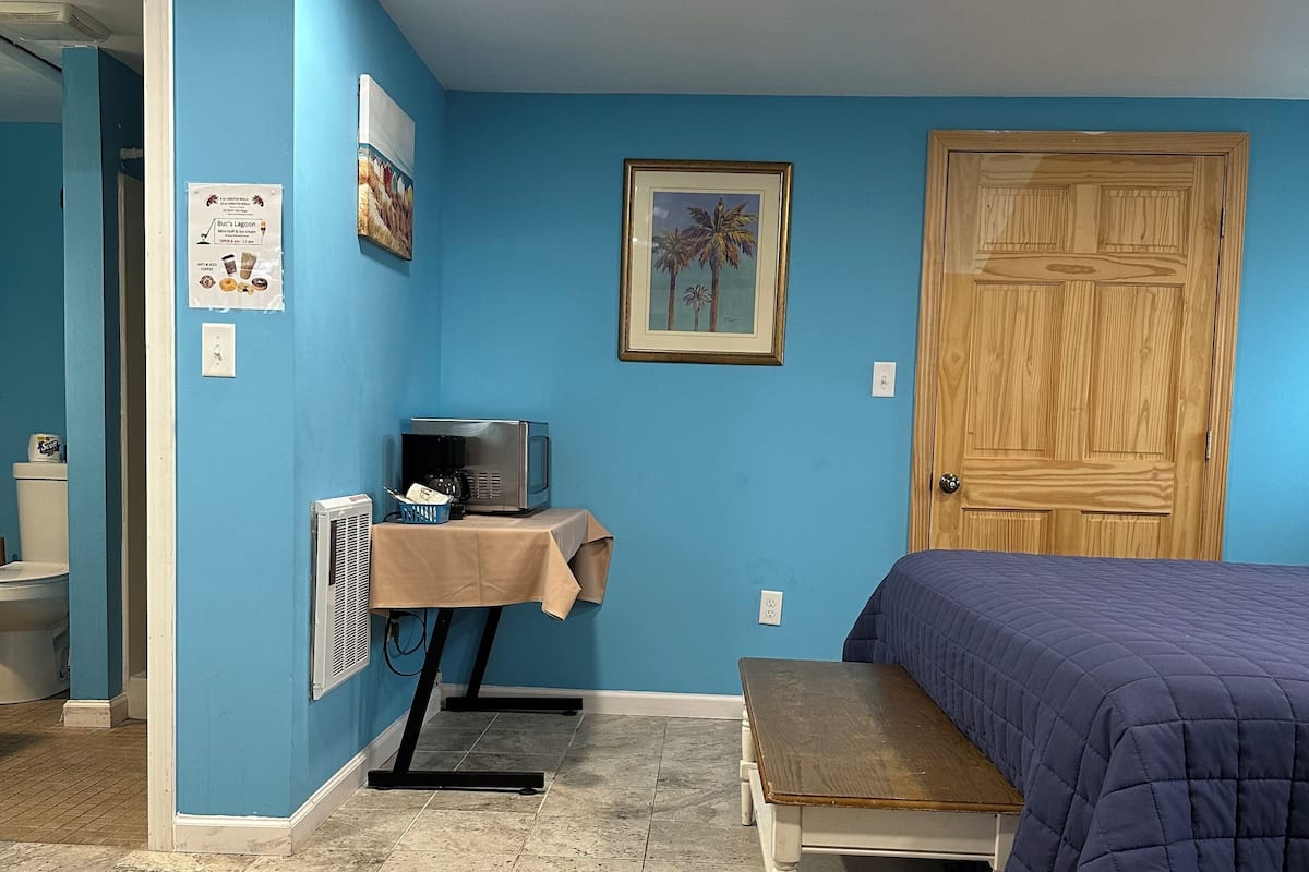 A room with blue walls, a wooden door, a bed with a blue blanket, a desk with a computer, and a framed picture on the wall.