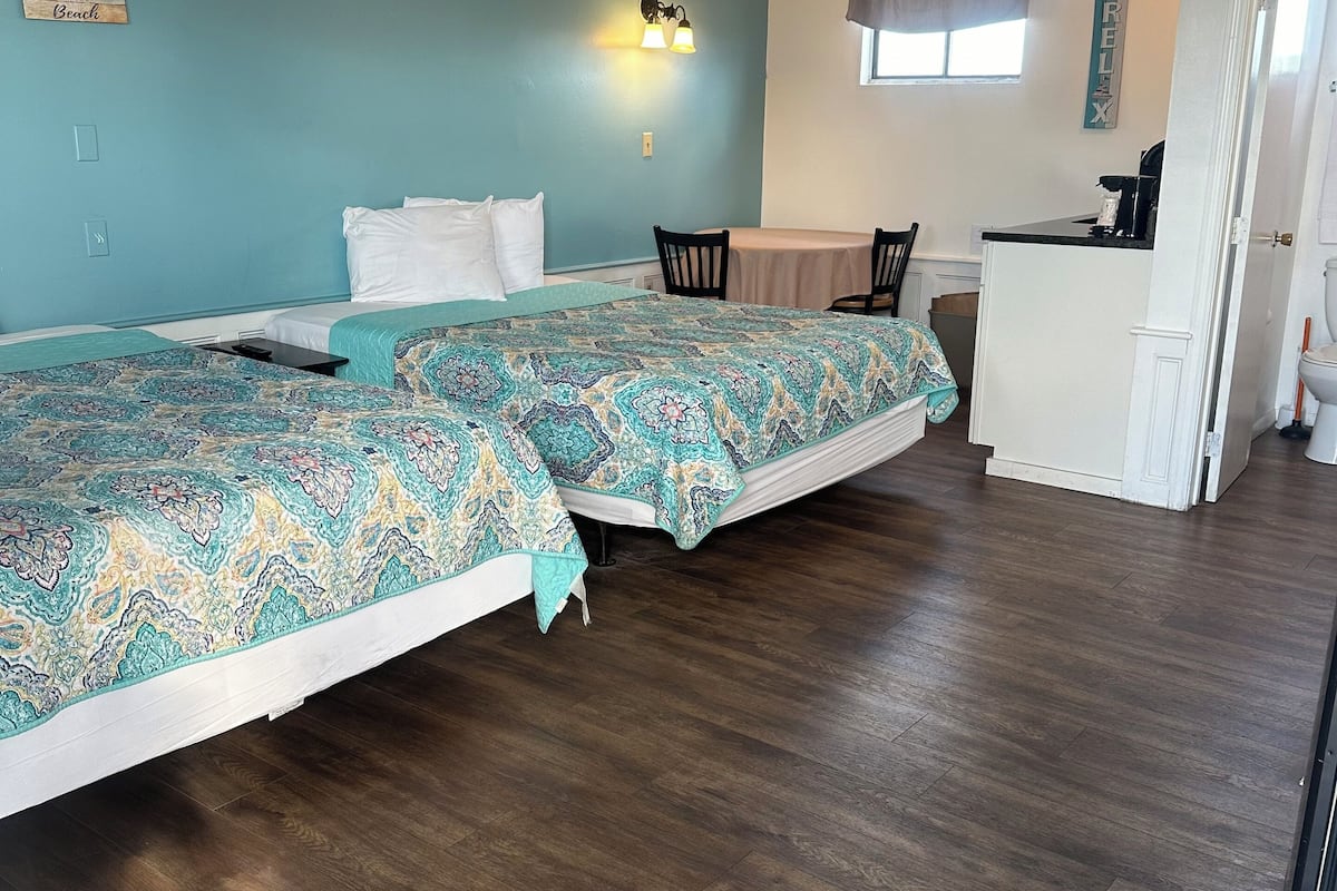 A room with two beds, a ceiling fan, a small kitchenette, and a window.