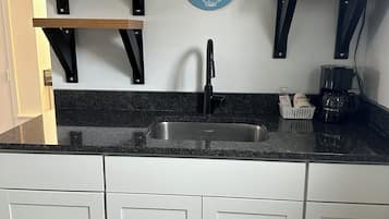 A kitchen with white cabinets, a black countertop, a stainless steel sink, wooden shelves, a microwave, and a decorative sign that reads "Seas the Day".
