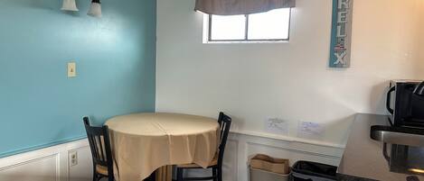 Eine kleine Küchenzeile mit Tisch und Stühlen, ein Fenster mit Vorhang sowie ein Schild an der Wand.