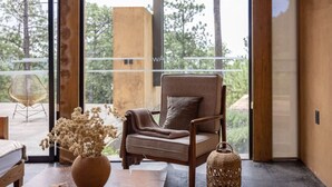Een moderne woonkamer met een groot raam, een comfortabele fauteuil, een klein tafeltje met een vaas gedroogde bloemen, en een geweven mand.