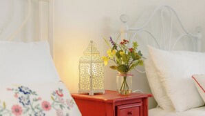 Un lit bien fait, avec du linge de lit blanc et un coussin à motifs floraux. Une table de chevet rouge porte une lanterne blanche et un vase rempli de fleurs.